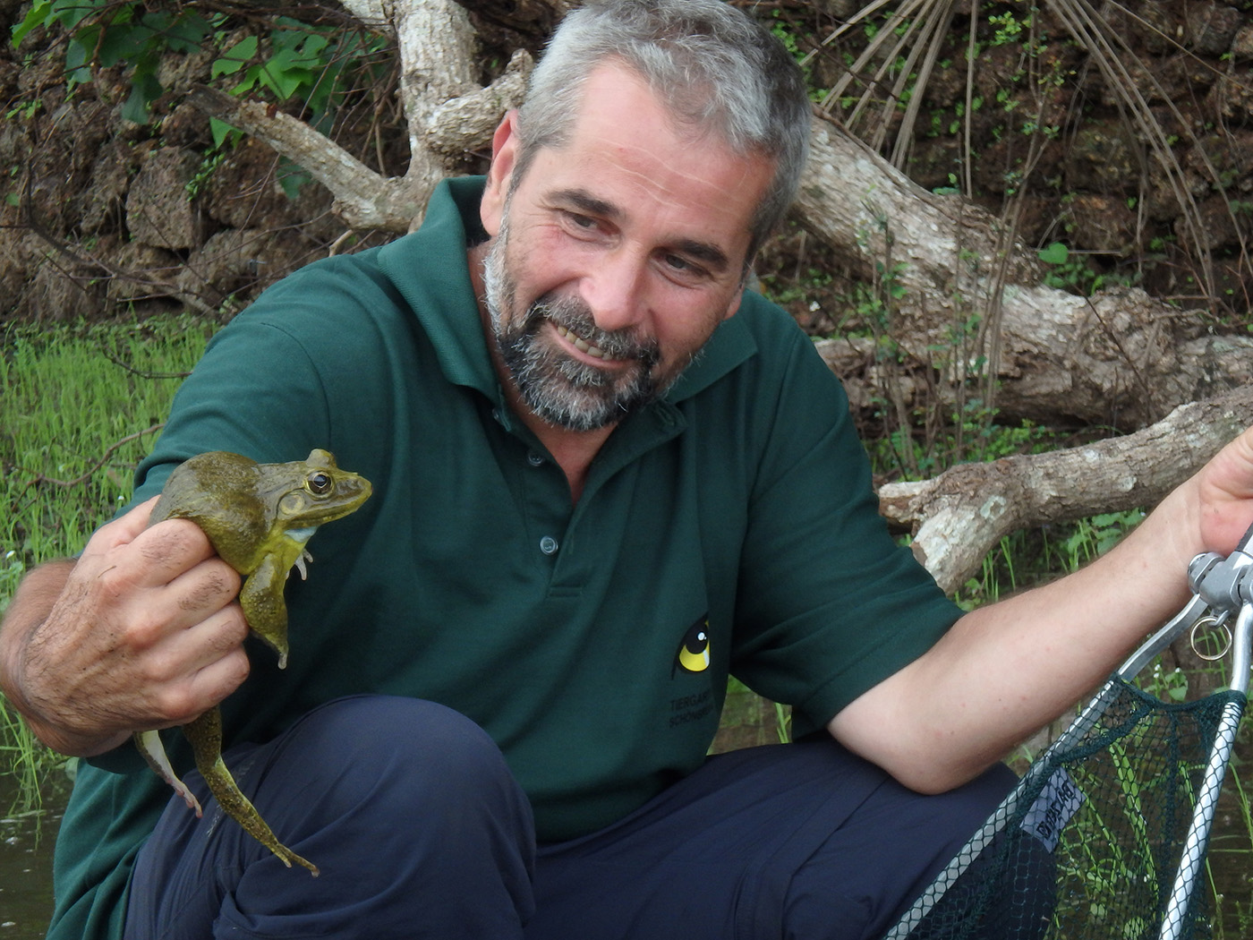 Ein Mann sitzt in der Natur auf dem Boden und hält einen Frosch in der Hand.